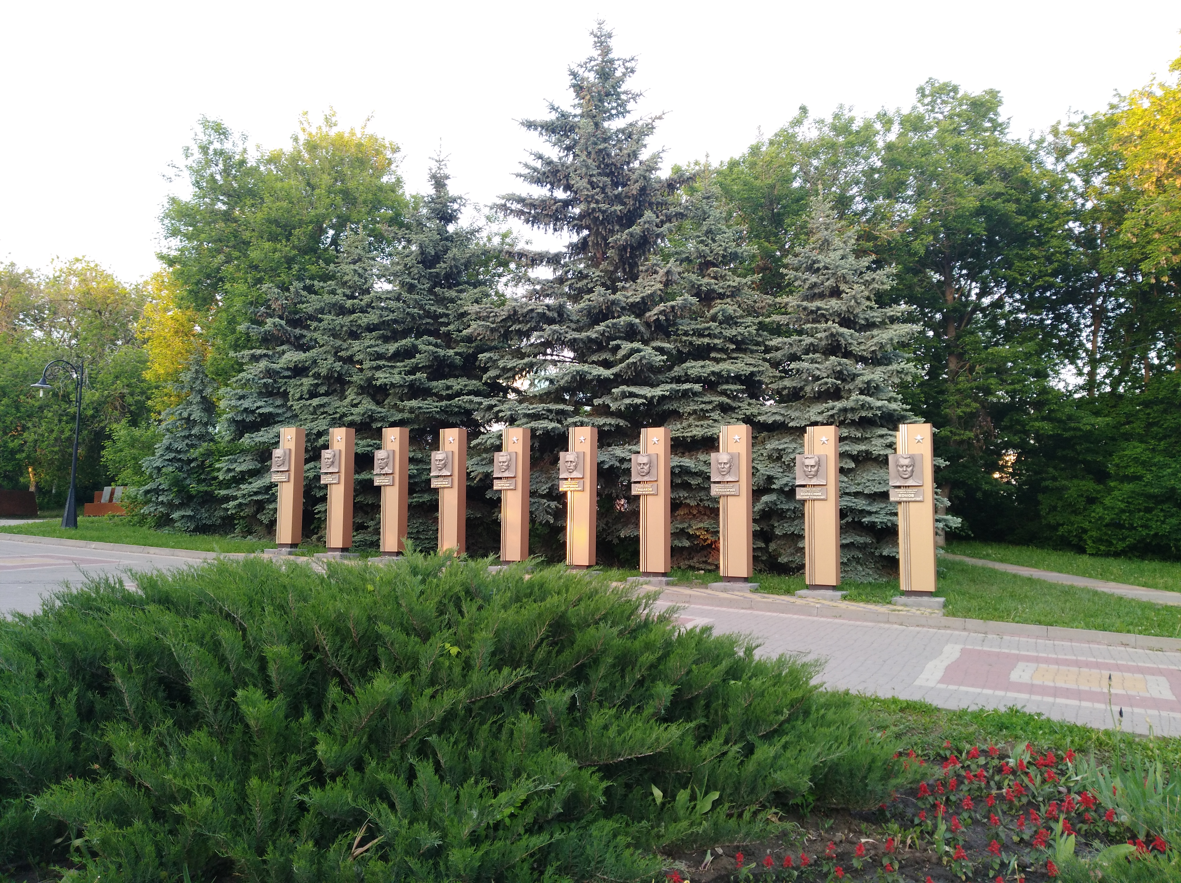 Ein Park mit einer Reihe von Bäumen in der Mitte, umgeben von blühenden Pflanzen, Gras, einem Weg, einem Straßenpfahl und einer Gruppe von Bäumen unter einem bewölkten Himmel, mit einem Gedenkschild für die Opfer von Tschernobyl.