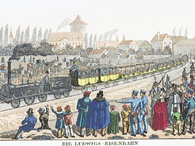 Eine Gruppe von Menschen steht um einen Zug auf einem Gleis, einige sitzen im Zug und andere stehen draußen, in der Nähe von Gebäuden und Bäumen, mit Text 'Ludwigs-Eisenbahn, die erste Bahnstation in Deutschland' unten.