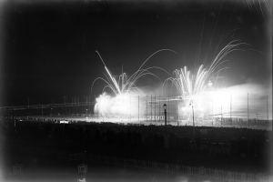 Ein Schwarz-Weiß-Foto einer Feuerwerksshow am Himmel, mit einer Brücke im Hintergrund, Laternen, die die Gegend beleuchten, und einem Zaun unten.