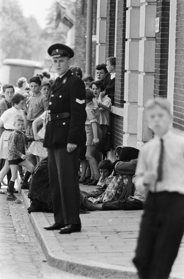 Ein Polizist steht vor einer Gruppe von Kindern, mit einem Gebäude auf der rechten Seite, Bäumen und Himmel im Hintergrund, auf einem Schwarz-Weiß-Bild.