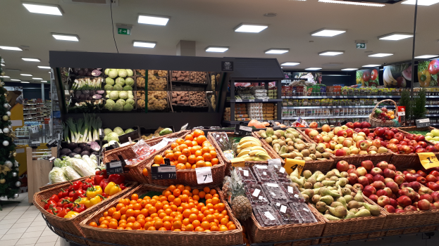 Ein Ladeneinrichtungsbild mit Körbüssern voller Obst und Gemüsse, Preisschildern, einem dekorierten Weihnachtsbaum links und Regalen mit anderen Artikeln im Hintergrund.