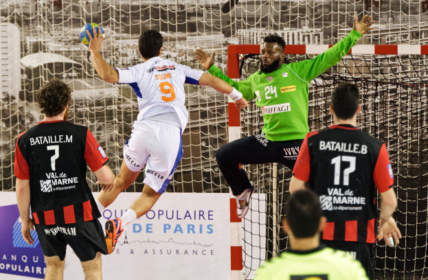 Eine Gruppe von Männern, die Handball spielen, wobei einer springt und den Ball hält, vor einem Torpfosten und einer Tafel mit der Aufschrift "Ligue 1 - Bordeaux vs Marseille."
