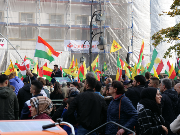 Eine große Gruppe von Menschen mit Fahnen und einer Person mit einem Mikrofon vor einem Gebäude, mit einem Fahrzeug, Baum, Pfosten, Lampen und Schildern mit Text, wahrscheinlich bei einer kurdischen Demonstration in Istanbul.