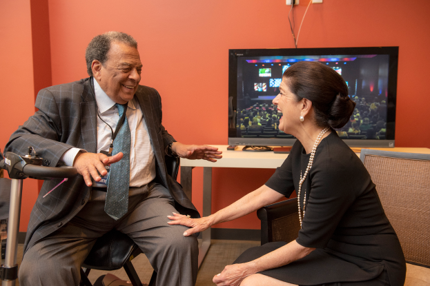 Ein Mann und eine Frau sitzen einander gegenüber auf Stühlen und unterhalten sich, mit einem Fernseher auf einem Tisch dazwischen, vor einer orangen Wand.