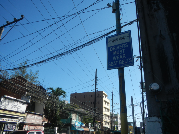 Eine Stadtstraße mit Autos, Strommasten mit Drähten, Gebäuden, Bäumen und Schildern, auf denen ein Schild mit der Aufschrift "Fahrer müssen den Sicherheitsgurt anlegen" zu sehen ist.