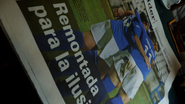 Eine Zeitung auf einem Tisch zeigt drei Männer in blauen T-Shirts, möglicherweise beim Fußballspielen, mit Text am unteren Bildrand.