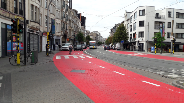 Eine Stadtstraße mit einer roten Fahrradspur, fahrende Fahrzeuge, Fußgänger auf dem Gehweg, Gebäude auf beiden Seiten, Bäume im Hintergrund und einen klaren blauen Himmel.