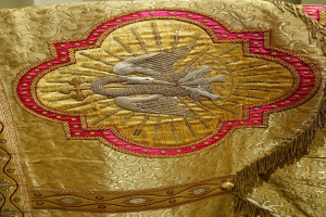 Nahaufnahme eines Priestergewandes mit goldener und roter Stickerei vor einer Wand als Hintergrund, mit detaillierter und luxuriöser Verarbeitung.