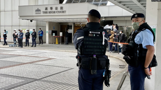 Zwei Polizisten in Uniform, mit Mützen und Masken, stehen vor einem Gebäude am Hong Kong International Airport, umgeben von einer Gruppe von Menschen, einige halten Kameras, mit Stangen im Hintergrund, die mit Bändern geschmückt sind.