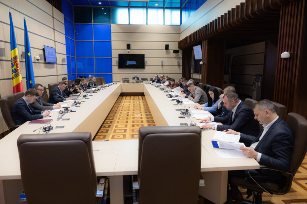 Eine Gruppe von Menschen sitzt um einen langen Tisch mit Papieren, Büchern, Mikrofonen und anderen Gegenständen während einer Sitzung des Europäischen Parlaments in Brüssel, mit Fahnen auf der linken Seite und Bildschirmen und Lichtern im Hintergrund.