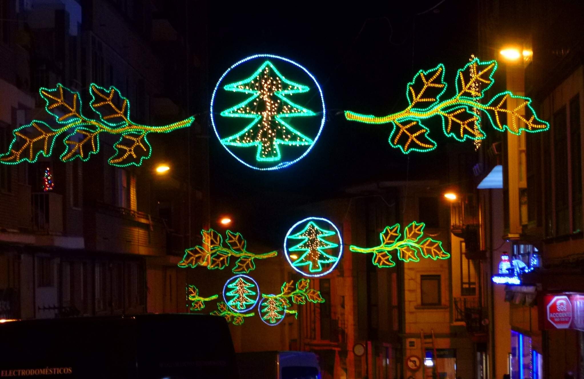 Eine nächtliche Stadtstraße, die mit festlichen Weihnachtslichtern in Form von Blättern geschmückt ist, mit Fahrzeugen auf der Straße und Gebäuden auf beiden Seiten.