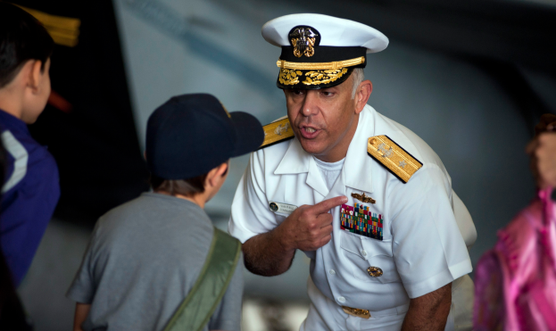 Ein Mann in wei├čer Milit├Ąruniform mit Abzeichen und M├╝tze spricht zu einer Gruppe von Kindern, mit unscharfem Hintergrund.