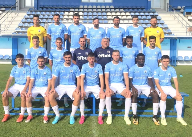 Eine Gruppe von Männern, einige sitzen auf Bänken und andere stehen auf einem Fußballfeld, feiern in einer Stadionatmosphäre mit Stühlen, Treppen, Geländern, Wänden und Fenstern im Hintergrund.