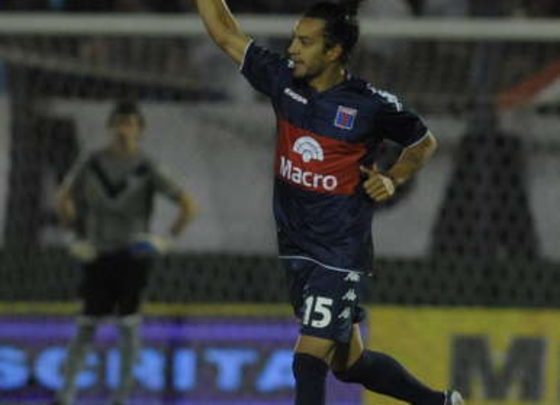 Ein Fußballspieler liegt auf dem Boden und feiert ein Tor, mit einer stehenden Person und einer Texttafel im Hintergrund, während eine jubelnde Menge unscharf in der Ferne zu sehen ist.