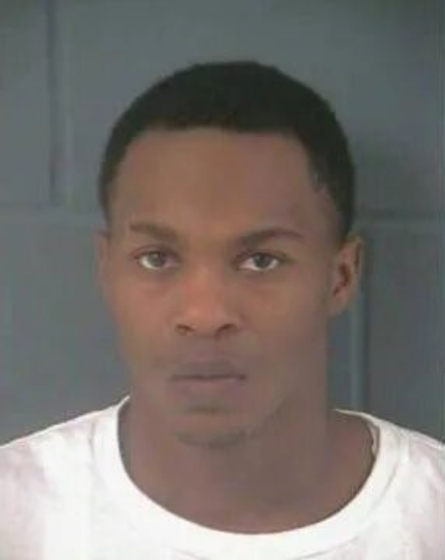 Ein Bild eines Mannes in einem weißen T-Shirt mit einer Wand im Hintergrund, der als Verdächtiger in einem Mordfall vermutet wird.