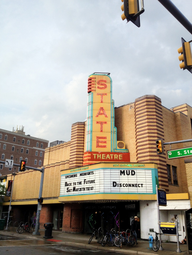 Das Staatstheater in Detroit, Michigan, mit einer Anzeigetafel, parkenden Fahrrädern, einer Person und umgebenden urbanen Elementen unter einem bewölktem Himmel.