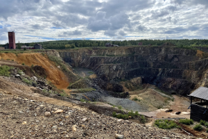 Großer Tagebau in einem Schmutzfeld umgeben von Bäumen, Gebäuden und einem Schuppen unter einem bewölkten Himmel, mit verstreuten Steinen und Pflanzen auf dem Boden.