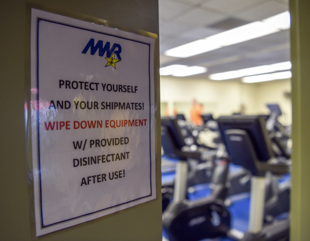 Ein Schild an einer Turnhalle mit der Aufschrift "Schützen Sie sich und Ihre Schiffskameraden" mit Laufbändern und Deckenbeleuchtung im Hintergrund, das Bild leicht unscharf, um Bewegung anzudeuten.