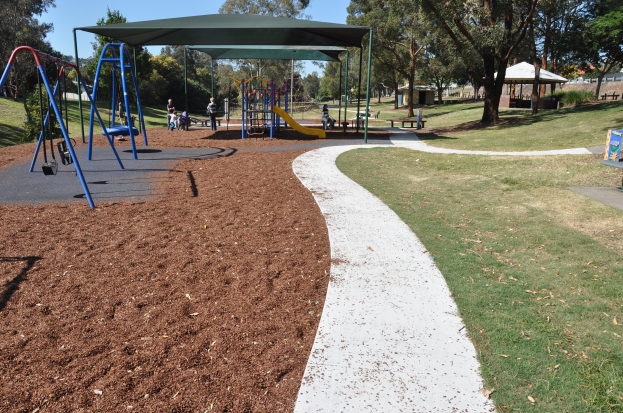 Ein Park mit einem Spielplatz mit Schaukeln, Rutschen und Bänken, umgeben von grünem Gras und Bäumen, mit Schuppen und einem klaren blauen Himmel im Hintergrund.