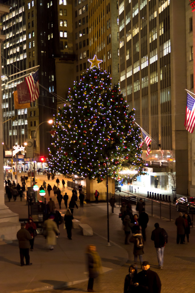 Ein Weihnachtsbaum, der mit Menschen geschmückt ist, mit einer Fahne auf einem Mast links, einer weiteren Fahne auf einem Mast rechts und beleuchteten Gebäuden im Hintergrund.