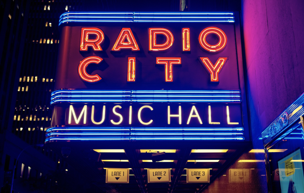 Außenansicht des Radio City Music Hall in New York City bei Nacht mit heller Beleuchtung und hohen umliegenden Gebäuden, einschließlich eines "Radio City"-Schilds und weiterer Schilder auf der rechten Seite.