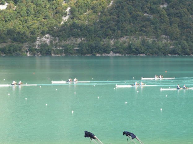 Eine Gruppe von Menschen, die in einem Wettbewerb auf einem Gewässer rudern, mit Bäumen und einem Hügel im Hintergrund und Kleidung auf Ständen am Wasserrand.