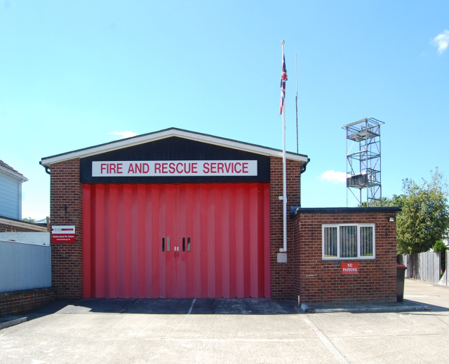 Feuerwehr- und Rettungsdienstgebäude mit roter Tür, Fenstern, einem Namensschild, einer Flaggenstange mit Flagge, einem Metallturm, einem Zaun, Bäumen und einem bewölkten Himmel.