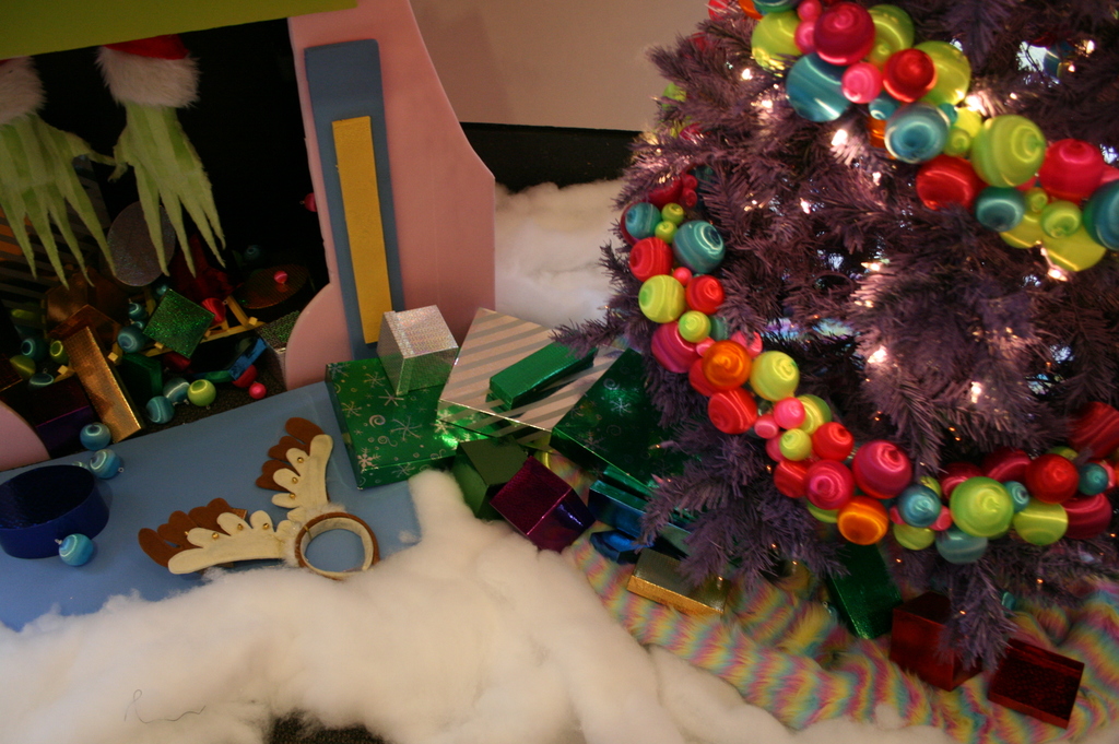 Eine Innenaufnahme mit einem geschmückten Weihnachtsbaum auf der rechten Seite, Geschenkboxen und Dekorationsgegenständen in der Mitte und einer Hütte mit Dekorationen oben links in der Ecke, mit Watte und Schafwolle unten.