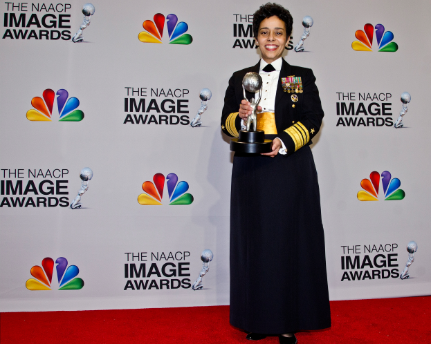 Eine Frau in militärischer Uniform steht auf einem roten Teppich, lächelt und hält eine Trophäe, mit einer Tafel im Hintergrund, die Logos und Text anzeigt, dass sie bei den NAACP Image Awards ist.