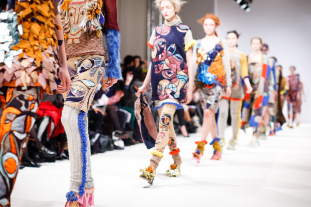 Eine Gruppe Von Frauen, Die Eine Laufsteg In London Fashion Week Mit Einer Zuschauersitzend Im Hintergrund Unter Helligen Lichtern Gehen.
