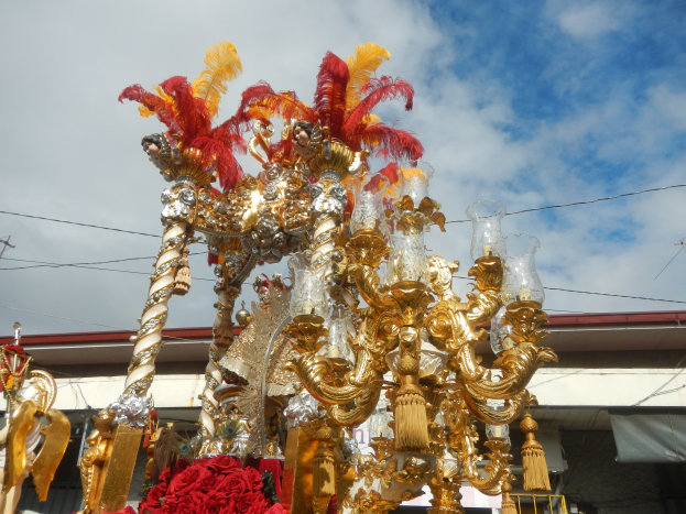 Ein großes goldenes und rotes Float, das mit Blumen und anderen Dekorationsgegenständen geschmückt ist, bei einem Karnevalsumzug, mit einem Gebäude, Strommasten mit Drähten und einem bewölkten Himmel im Hintergrund.