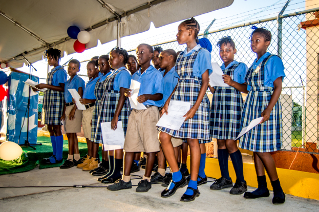 Schüler in Uniformen stehen auf einem Weg und halten Papiere, wobei ein Mädchen in ein Mikrofon auf einem Ständer spricht; dahinter ist eine Umzäunung mit einem Dach, das mit Ballons geschmückt ist.