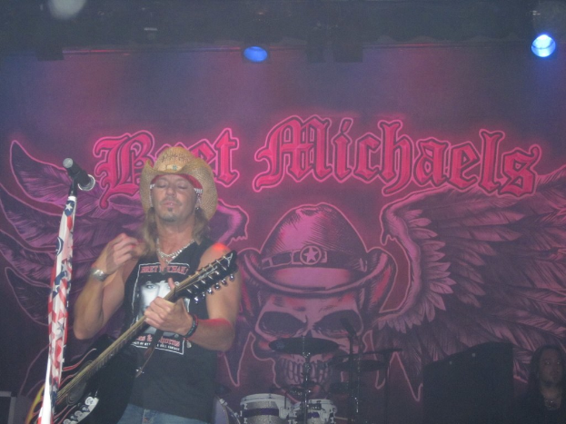 Eine Musikkonzertszene mit einem Gitarristen auf der Bühne, einer Person in der unteren rechten Ecke, einem Banner im Hintergrund und Drogen in der Mitte unten, mit Beleuchtung von oben.