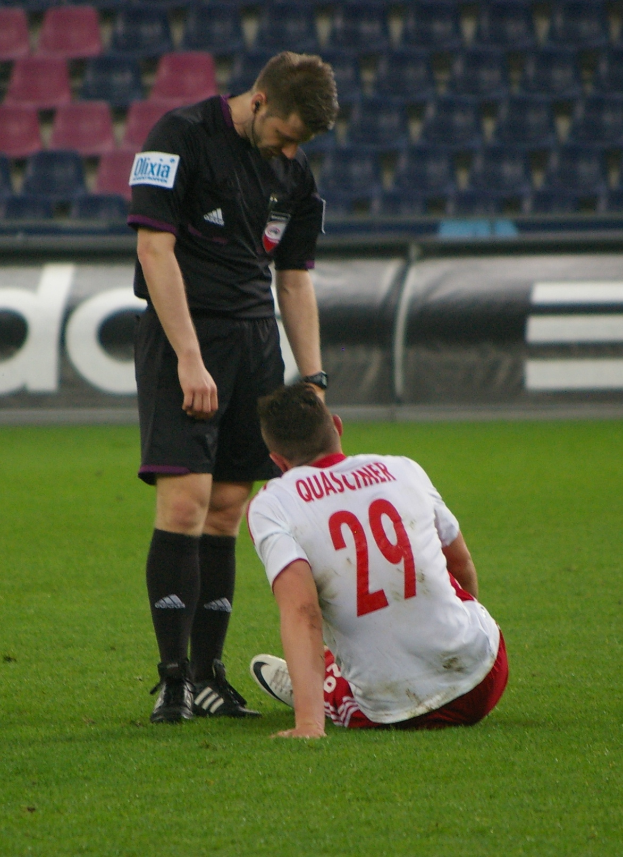 Ein Fussballspieler sitzt neben einem Schiedsrichter auf dem Boden in einem Stadion, beide tragen Sportkleidung, mit Tafeln und Stühlen im Hintergrund.