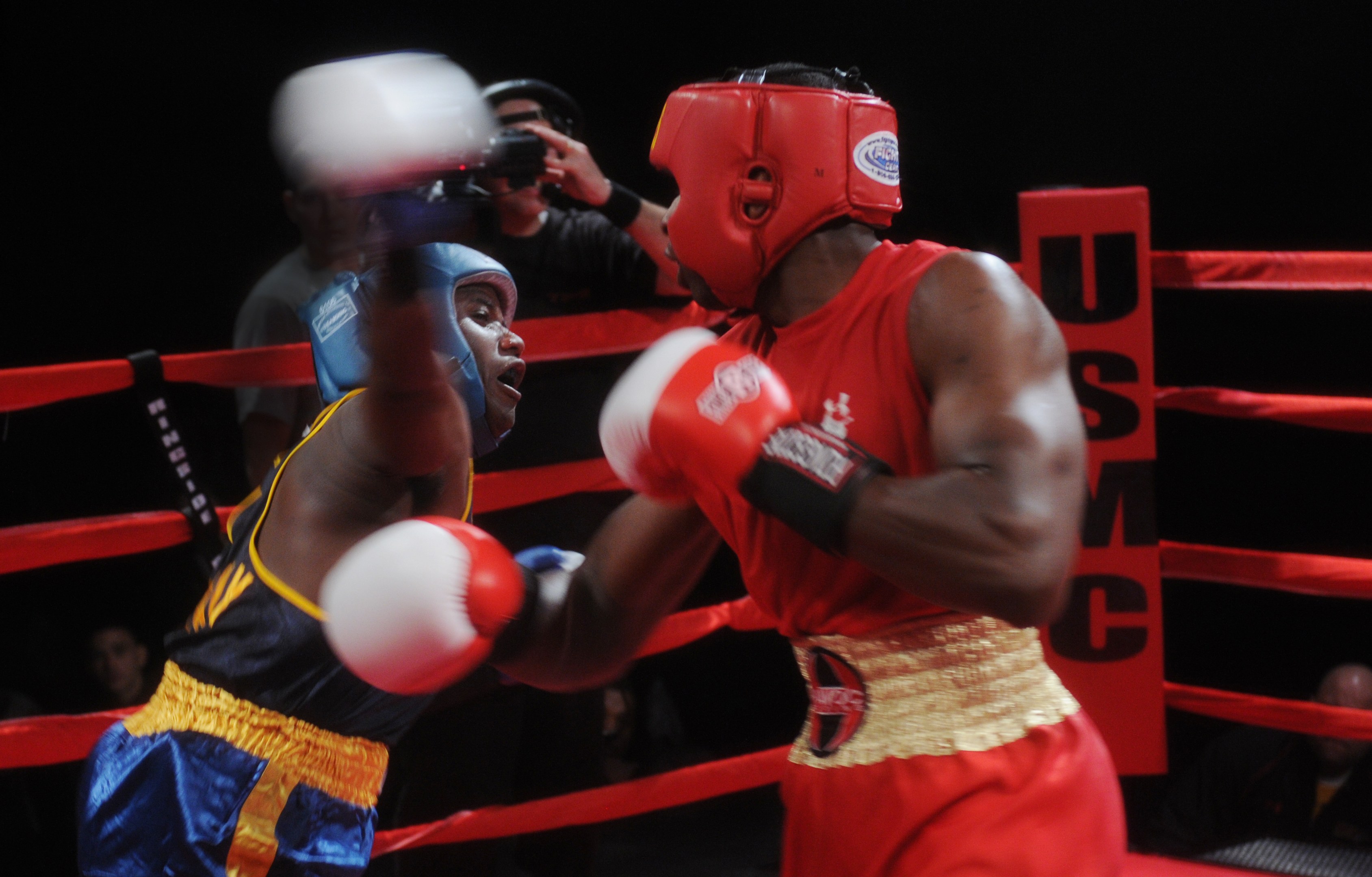Zwei Boxer im Ring bei einem Kampf, mit einem Fotografen und Zuschauern im Hintergrund bei dramatischer Beleuchtung.