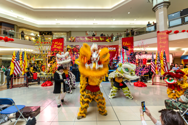 Ein lebhaftes chinesisches Neujahrsfest in einem Einkaufszentrum mit tanzenden Menschen in Kostümen und anderen, die auf Stühlen sitzen und mobile Telefone halten, umgeben von Flaggen, Bannern, Dekorationen und Lichtern.