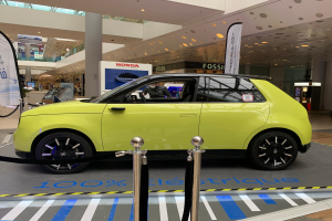 Ein gelbes wasserstoffbetriebenes Elektroauto, das drinnen auf einem Parkplatz steht, umgeben von Absperrpollern mit Seilen, Bannern und Schildern, mit einer Person im Auto und einer Gruppe von Menschen in der Nähe.