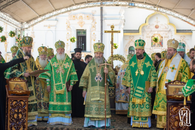 Gruppe von Priestern vor einer Kirche während einer religiösen Zeremonie, mit einer Person, die ein Buch und ein Mikrofon hält, links, und einem Kreuzsymbol, Blumen und einem Gebäude im Hintergrund.