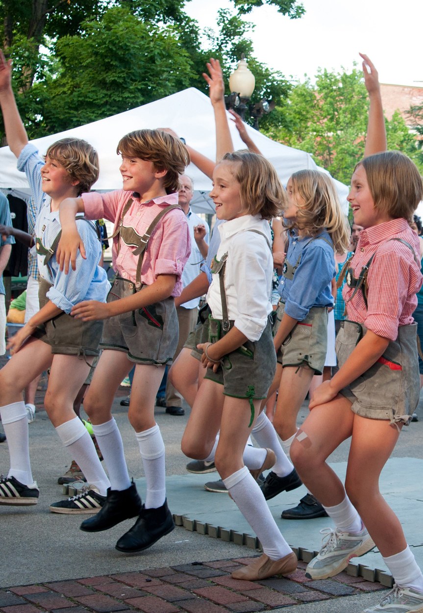 Kinder in traditioneller bayrischer Tracht tanzen auf einer Straße, umgeben von einer Menge, Zelten, Laternenmasten, Bäumen, Gebäuden und einem klaren blauen Himmel.