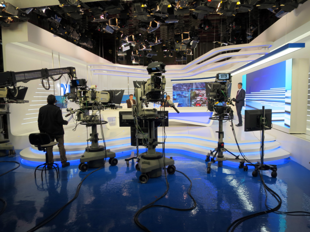 Ein TV-Studio mit mehreren Kameras auf dem Boden, einer sitzenden Person im Vordergrund und einigen stehenden Personen im Hintergrund, beleuchtet von Deckenlampen für eine Live-Übertragung.