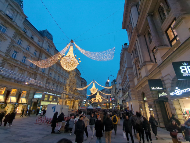 Eine belebte Stadtstraße voller Menschen, die unter festlichen Weihnachtslichtern spazieren gehen, gesäumt von Gebäuden mit Fenstern, unter einem klaren blauen Himmel.