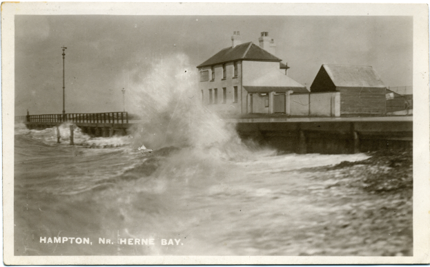 Ein Schwarz-Weiß-Foto einer großen Welle, die gegen eine Mole kracht, mit Häusern, Pfählen und einer Brücke im Hintergrund und Text am unteren Rand.