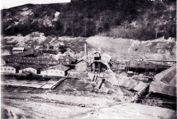 Schwarzes und wei├čes Foto eines Kohlebergbaus mit Geb├Ąuden, Masten und B├Ąumen im Hintergrund und Text am unteren Rand.