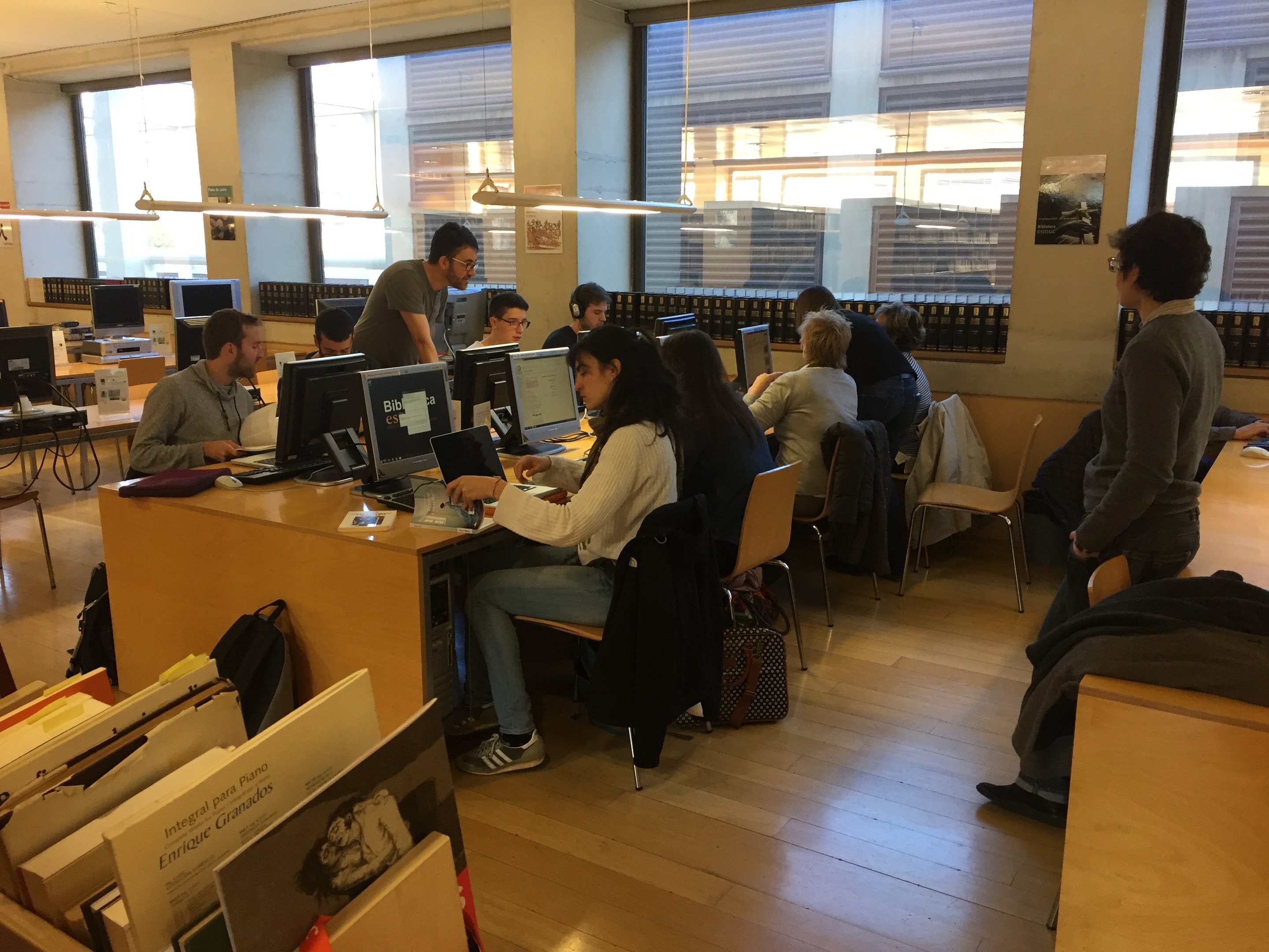 Gruppe von Menschen, die an Tischen in einer Bibliothek arbeiten, umgeben von Bücherregalen, Monitoren, Tastaturen, Mäusen, Taschen auf dem Boden, Deckenlampen und Glasfenstern im Hintergrund.