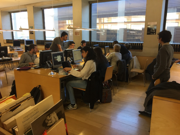 Gruppe von Menschen, die an Tischen in einer Bibliothek arbeiten, umgeben von Bücherregalen, Monitoren, Tastaturen, Mäusen, Taschen auf dem Boden, Deckenlampen und Glasfenstern im Hintergrund.