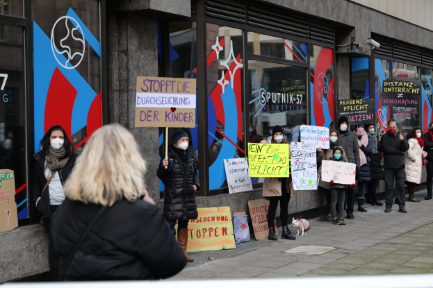 Eine Gruppe von Menschen mit Masken und Schildern protestiert vor einem Gebäude mit Glaswänden und -türen, begleitet von Texttafeln, Kameras und einem Hund.