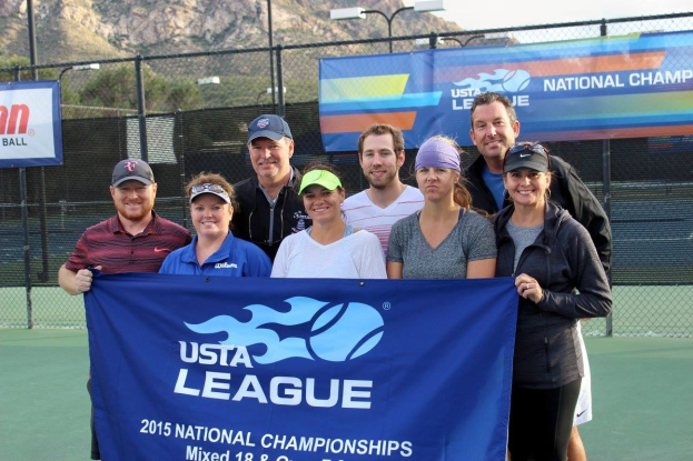 Gruppe von Menschen auf einem Tennisplatz mit einem "2015 USTA National Championships"-Banner, einem Zaun, Bannern, Laternenmasten, Bäumen, Bergen und einem klaren blauen Himmel im Hintergrund.