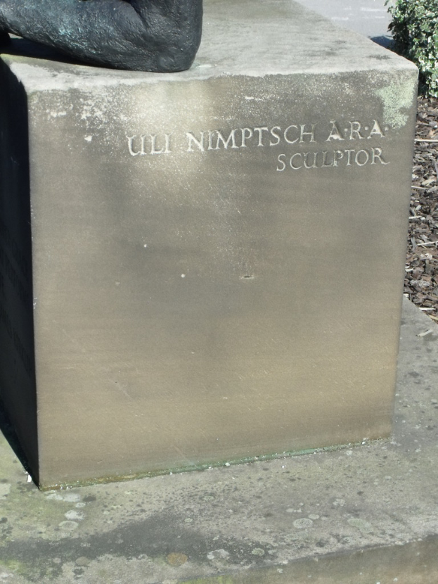 Ein Stein mit Text in der Vordergrund, möglicherweise eine Statue oben, mit trockenen Zweigen, Erde und Pflanzen rechts und einer Straße im Hintergrund.