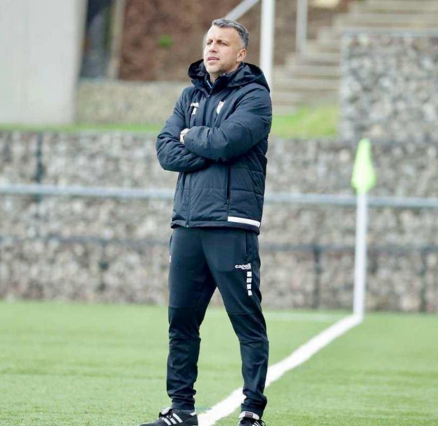 Ein Mann in schwarzer Jacke und Hose steht auf einem Fussballfeld mit überkreuzten Armen, der scheinbar ein Trainer wahrend einer Trainingseinheit ist.
