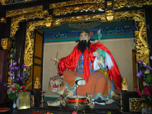 Eine Statue eines chinesischen Mönchs, der mit farbenfroher Stoff behangen ist, sitzt auf einer Tigerstatue innerhalb eines Bogens mit goldenen Verzierungen, vor ihm ein Blumenstrauß und in der Mitte eine Schüssel.